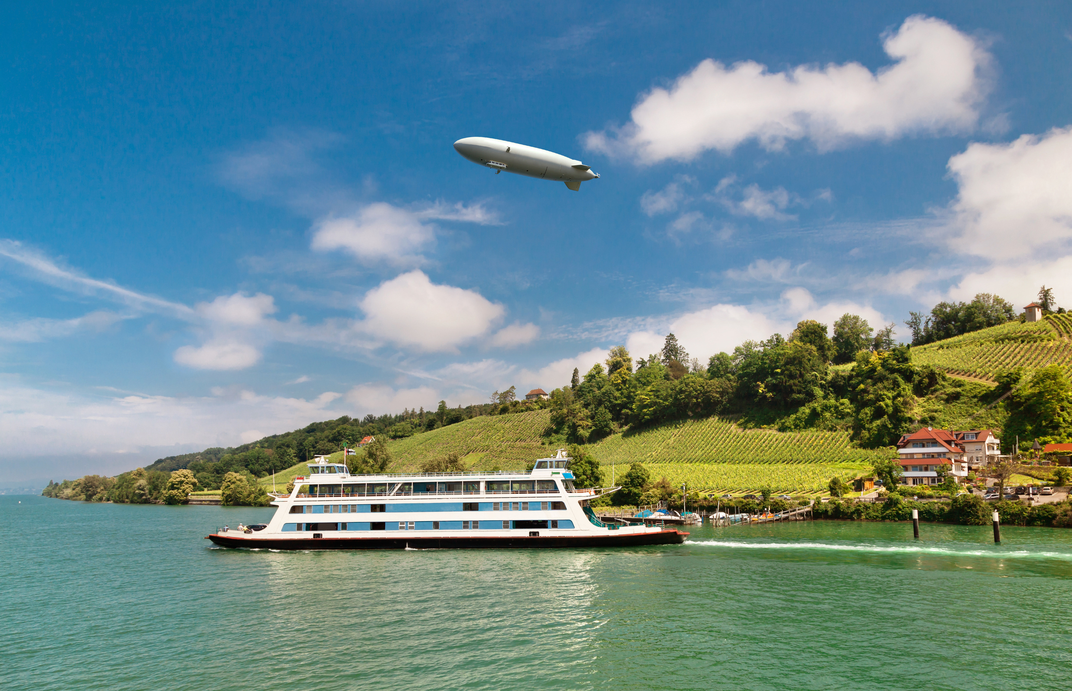 Ein Zeppelin schwebt über einer Flusslandschaft mit Weinbergen und einer Fähre.