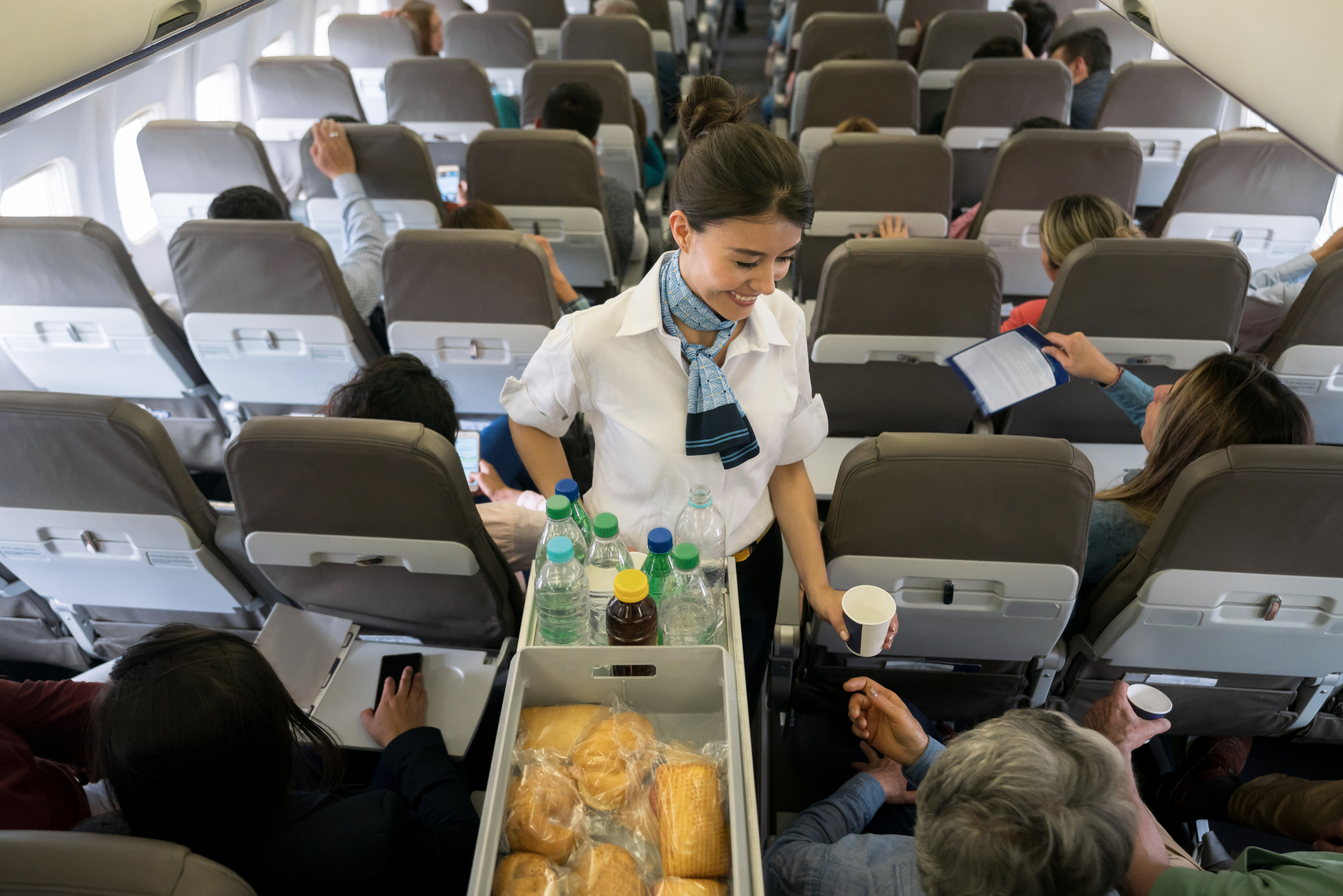 Bruinharige glimlachende stewardess met blauwe sjaal deelt eten en drinken uit aan passagiers op een vliegtuig