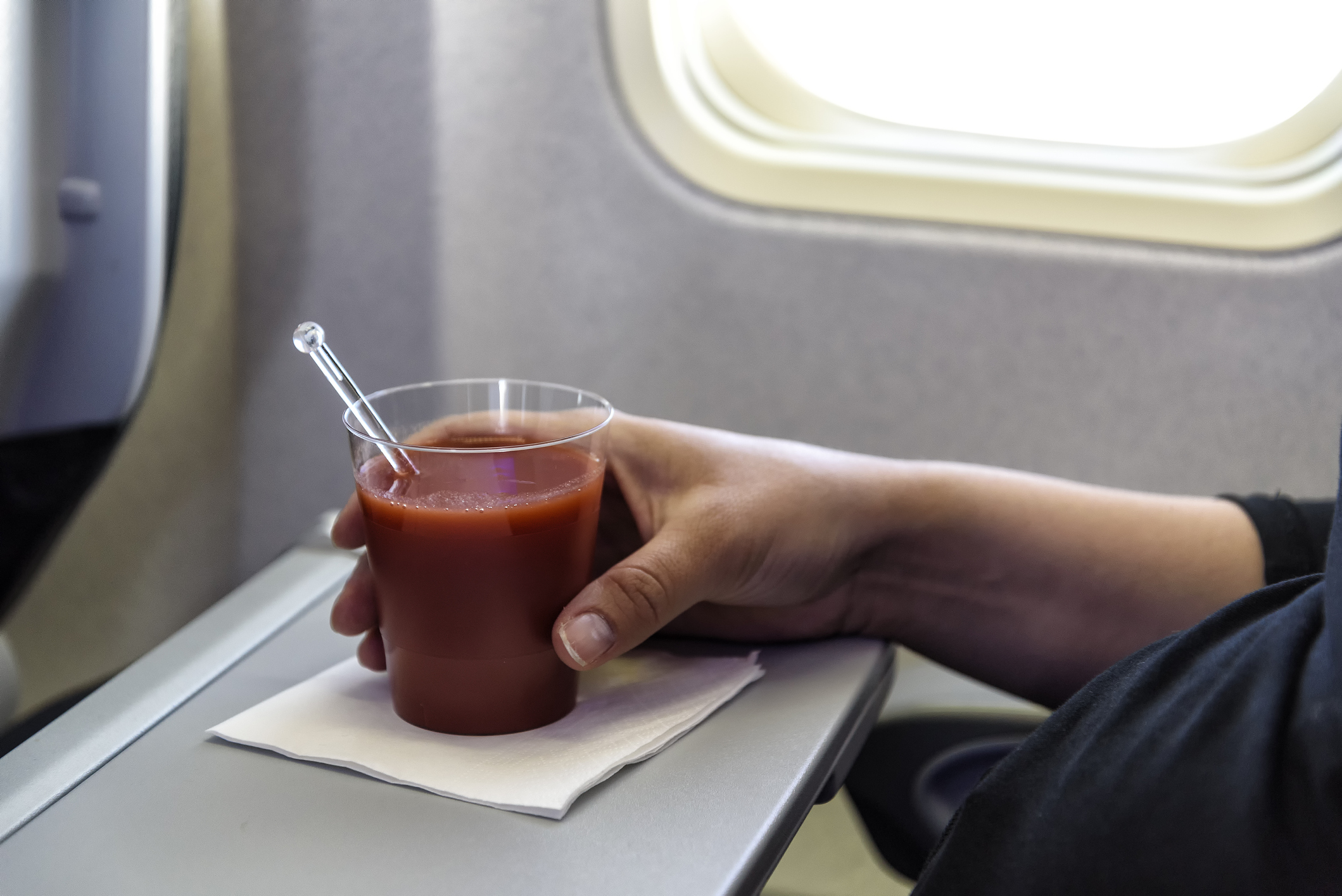 Nahaufnahme einer Hand, die einen Plastikbecher mit Tomatensaft umfasst, der auf einer Serviette eines Klapptisches im Flugzeug steht