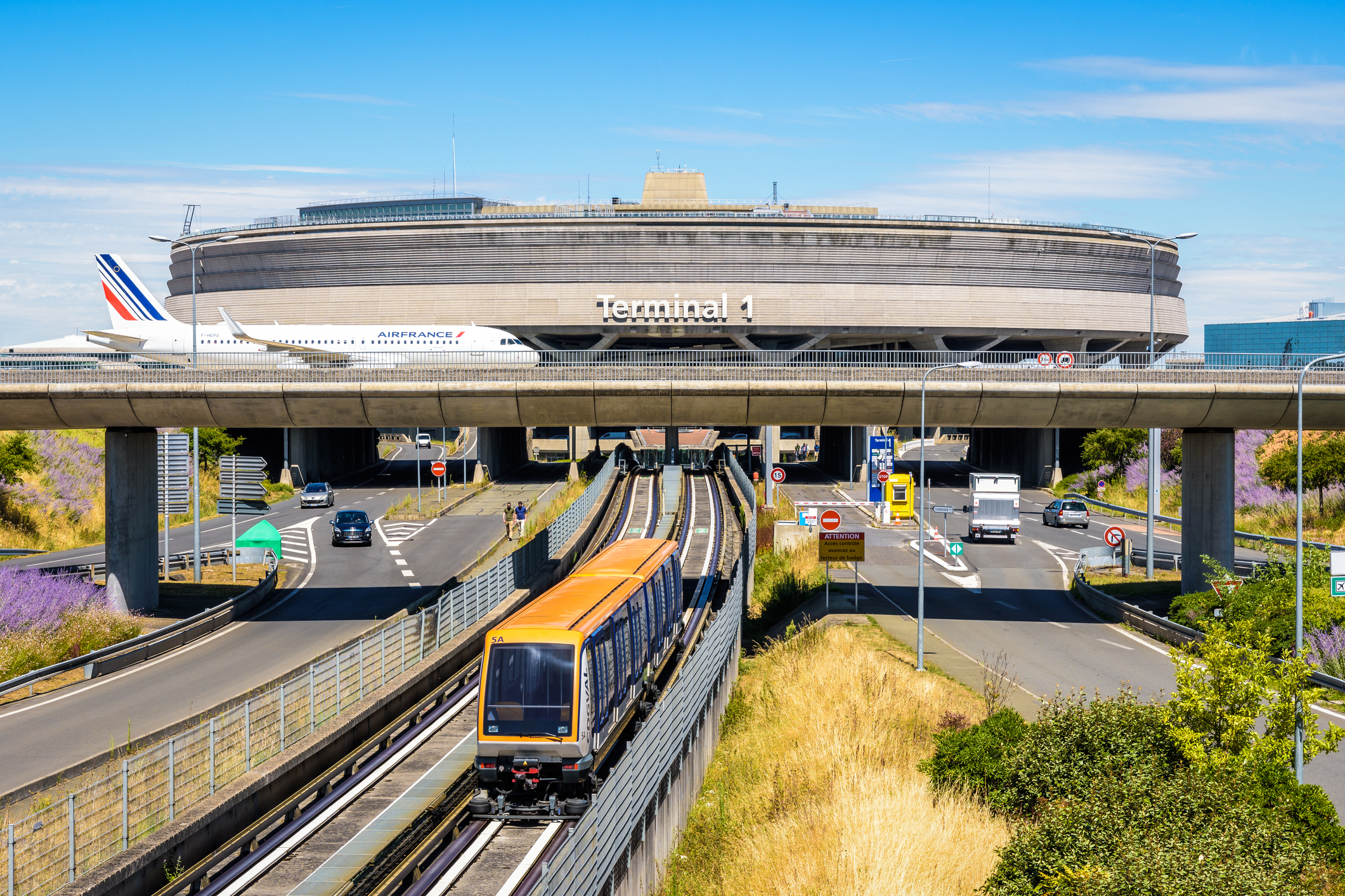 Ein Bild von Terminal 1 des Flughafens Charles de Gaulle, im Vordergrund ist eine Straßenbahn zu sehen, im Hintergrund überquert eine Air France Maschine auf einer Brücke eine Autobahn.