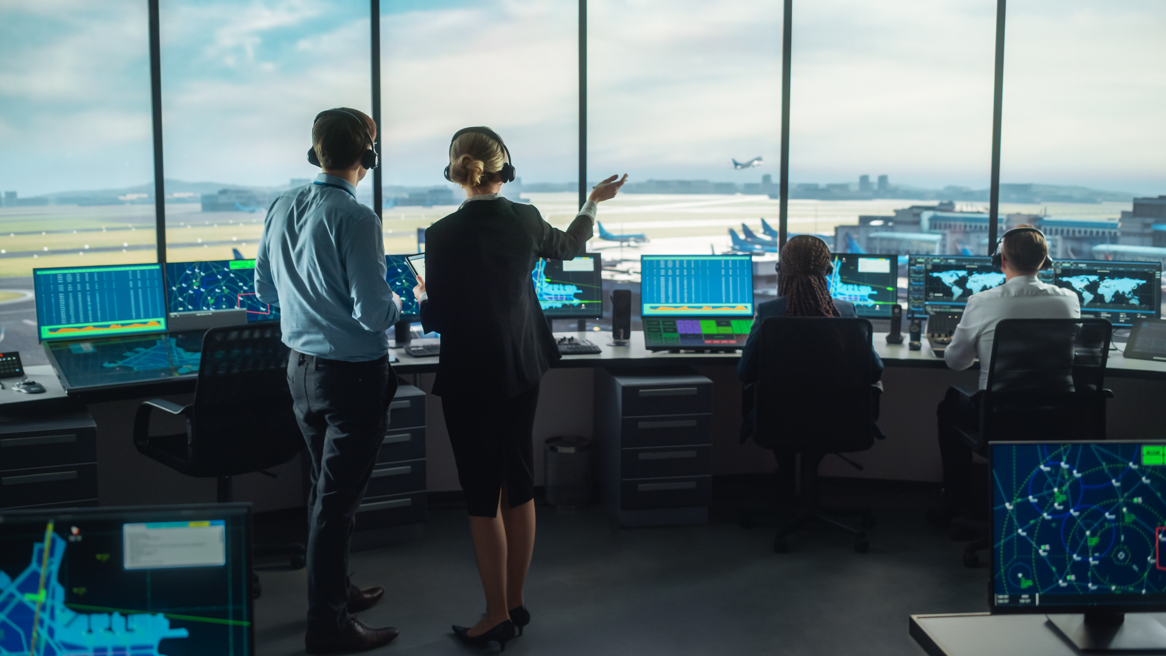 Im Inneren eines Airport-Towers befinden sich mehrere Fluglotsen und einige Bildschirme, im Hintergrund sind durch die Fensterfront Start- und Landebahnen und ein abhebendes Flugzeug zu sehen.