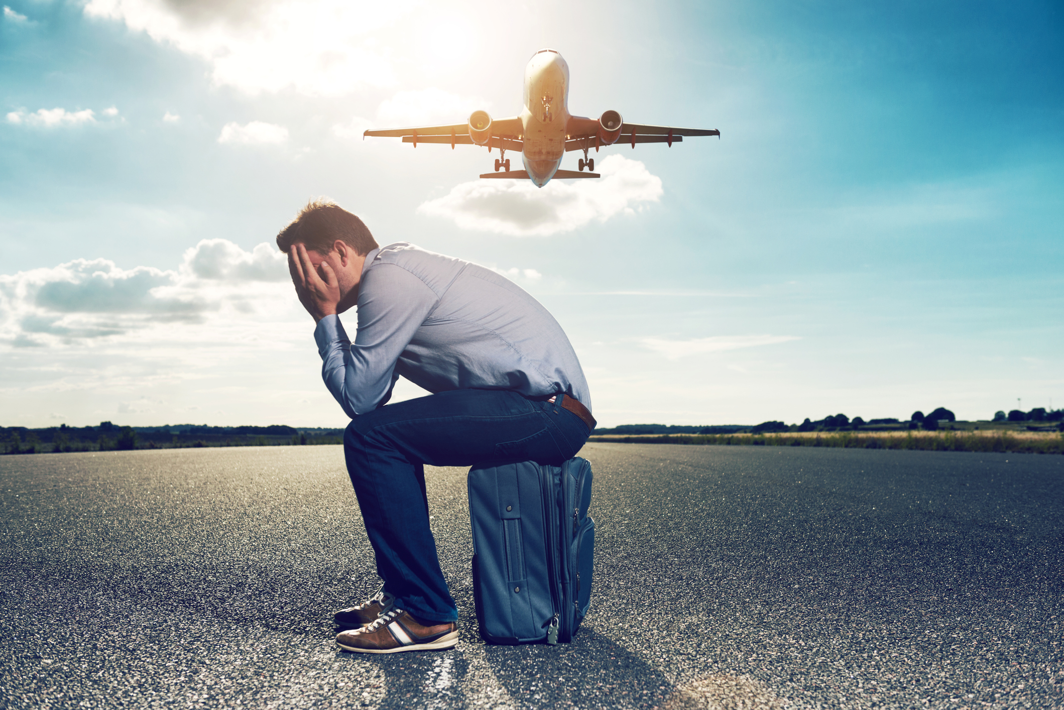 Een man zit op een blauwe koffer met zijn handen voor zijn gezicht terwijl achter hem een vliegtuig vliegt.