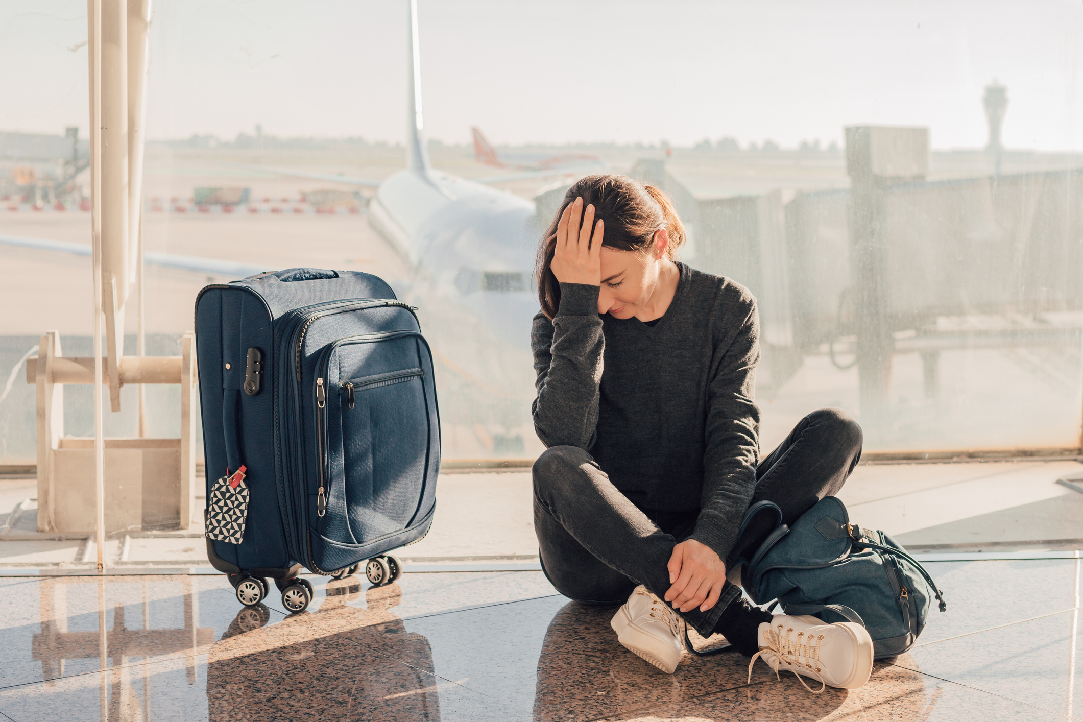 Eine Frau, die im Flughafen neben ihrem Koffer und mit ihrem Rucksack auf dem Boden sitzt, fasst sich an Kopf.
