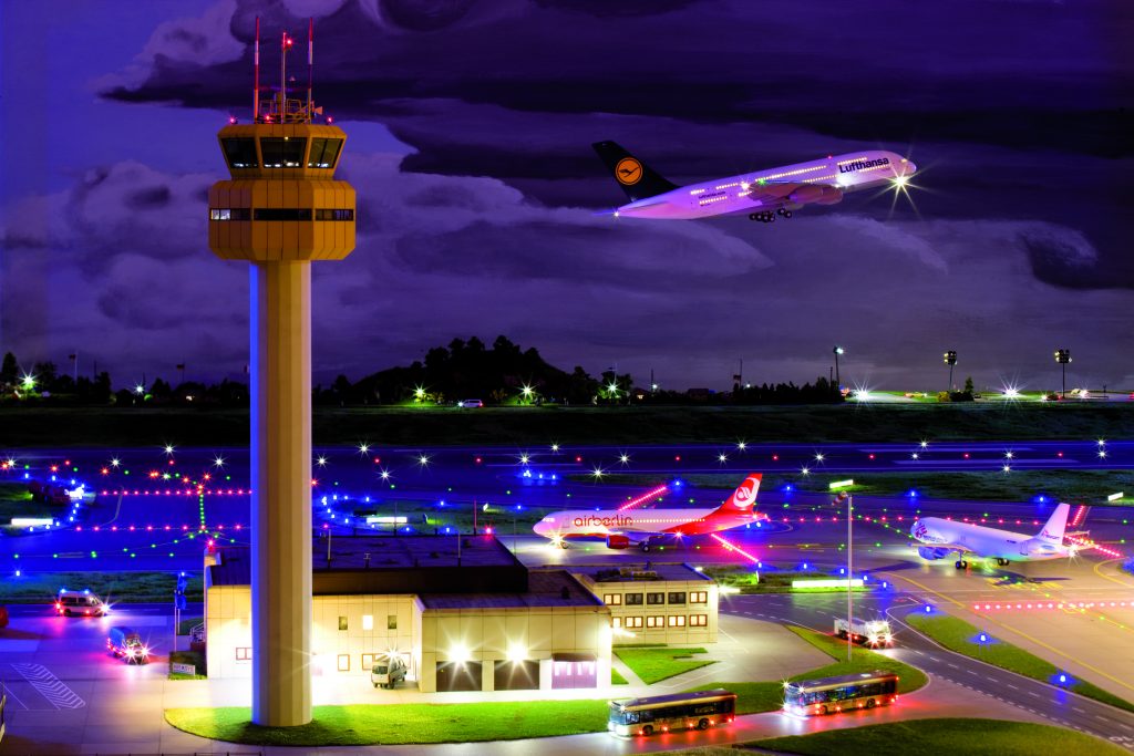Der Tower von Knuffingen Airport bei Nacht, im Hintergrund hebt eine Lufthansa-Maschine ab, zwei weitere Flugzeuge fahren zum Rollfeld.