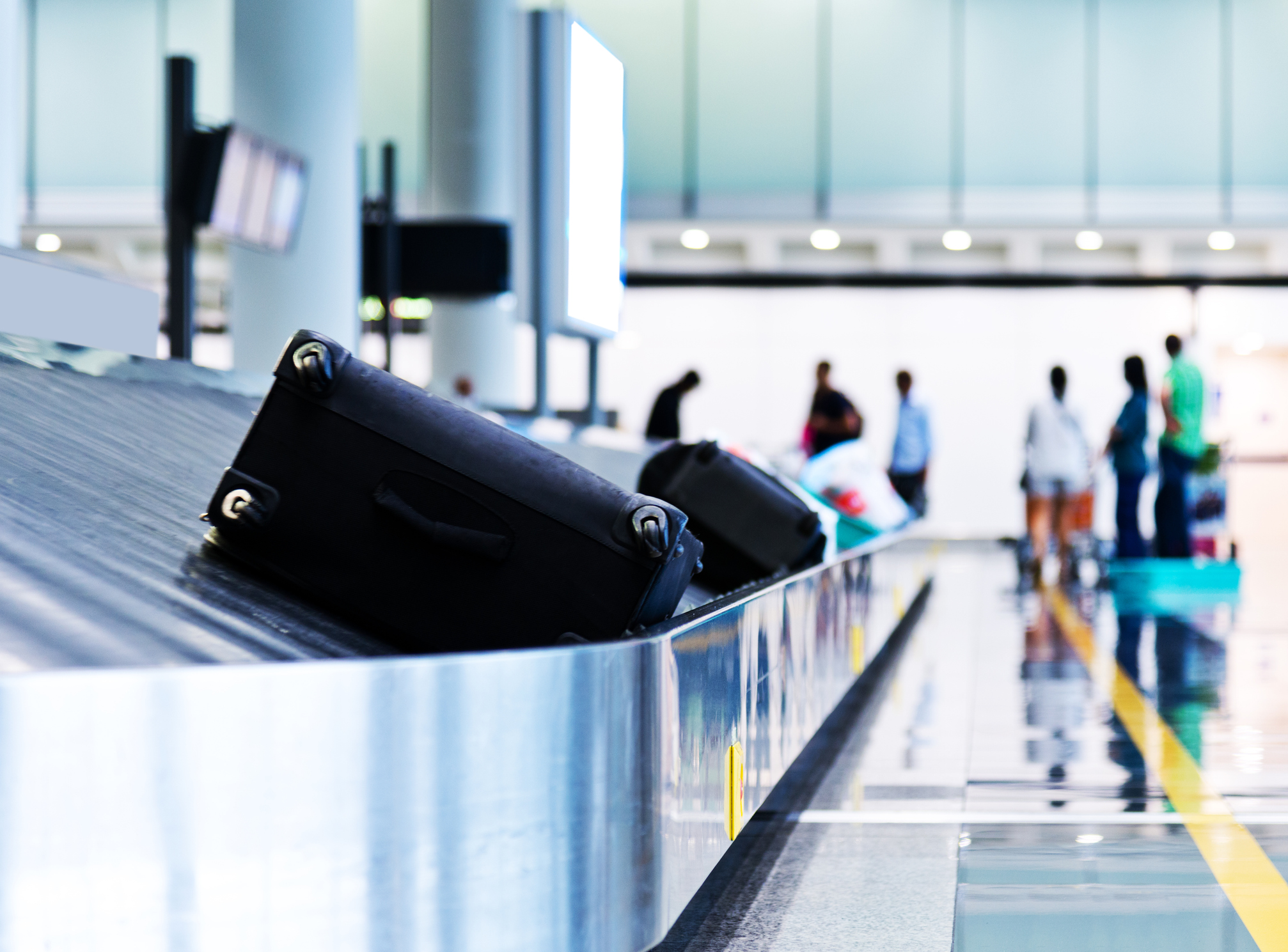 Bagagebånd i en lufthavn med to sorte kufferter i forgrunden og flere ventende personer i baggrunden