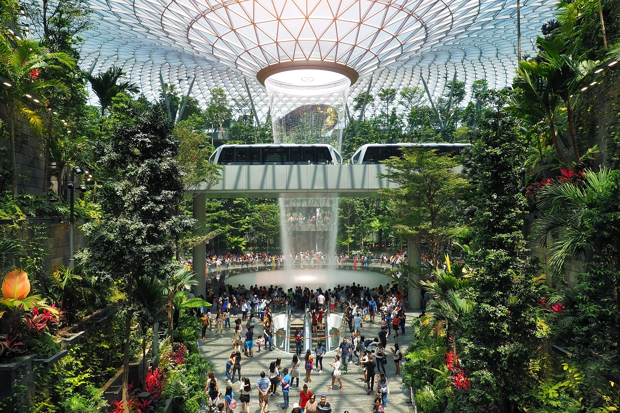 Eine Panorama-Aufnahme aus dem Inneren des Jewel Changi, zu sehen sind der Rain Vortex Wasserfall, tropische Pflanzen und viele Reisende.
