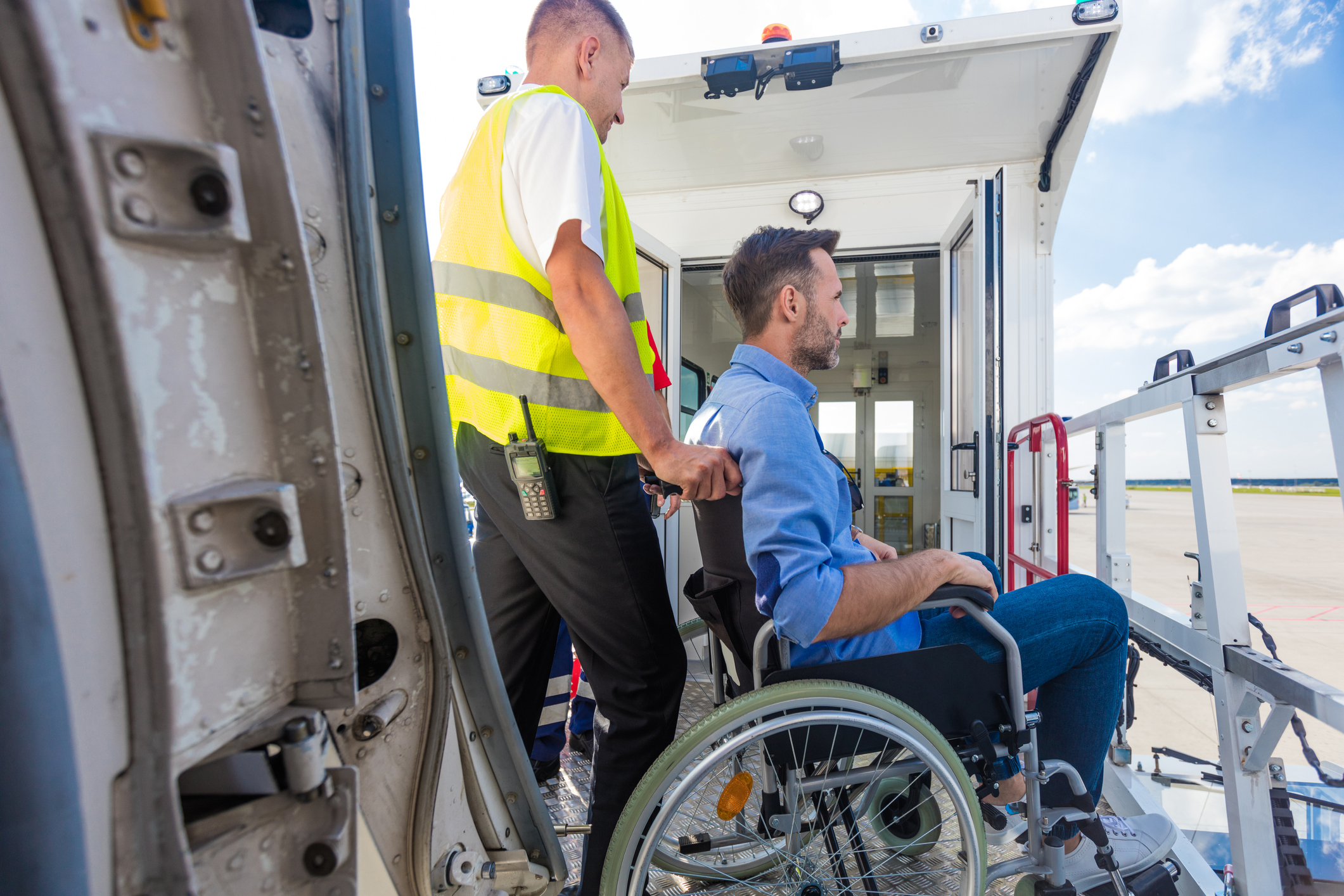 Ein Mann in einem Rollstuhl wird von einem Airline-Mitarbeiter geschoben, im Vordergrund ist die Flugzeugtür zu sehen, um Hintergrund die Gangway.