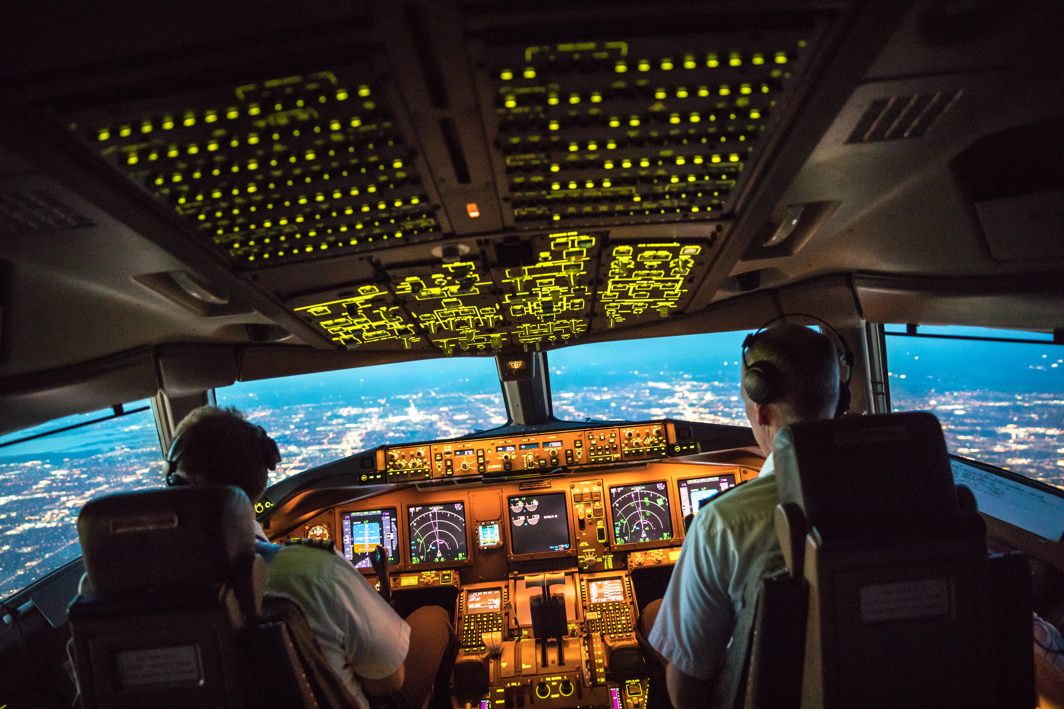 Ein beleuchtetes Flugzeugcockpit mit zwei Piloten beim Landeanflug auf eine Stadt am Abend.