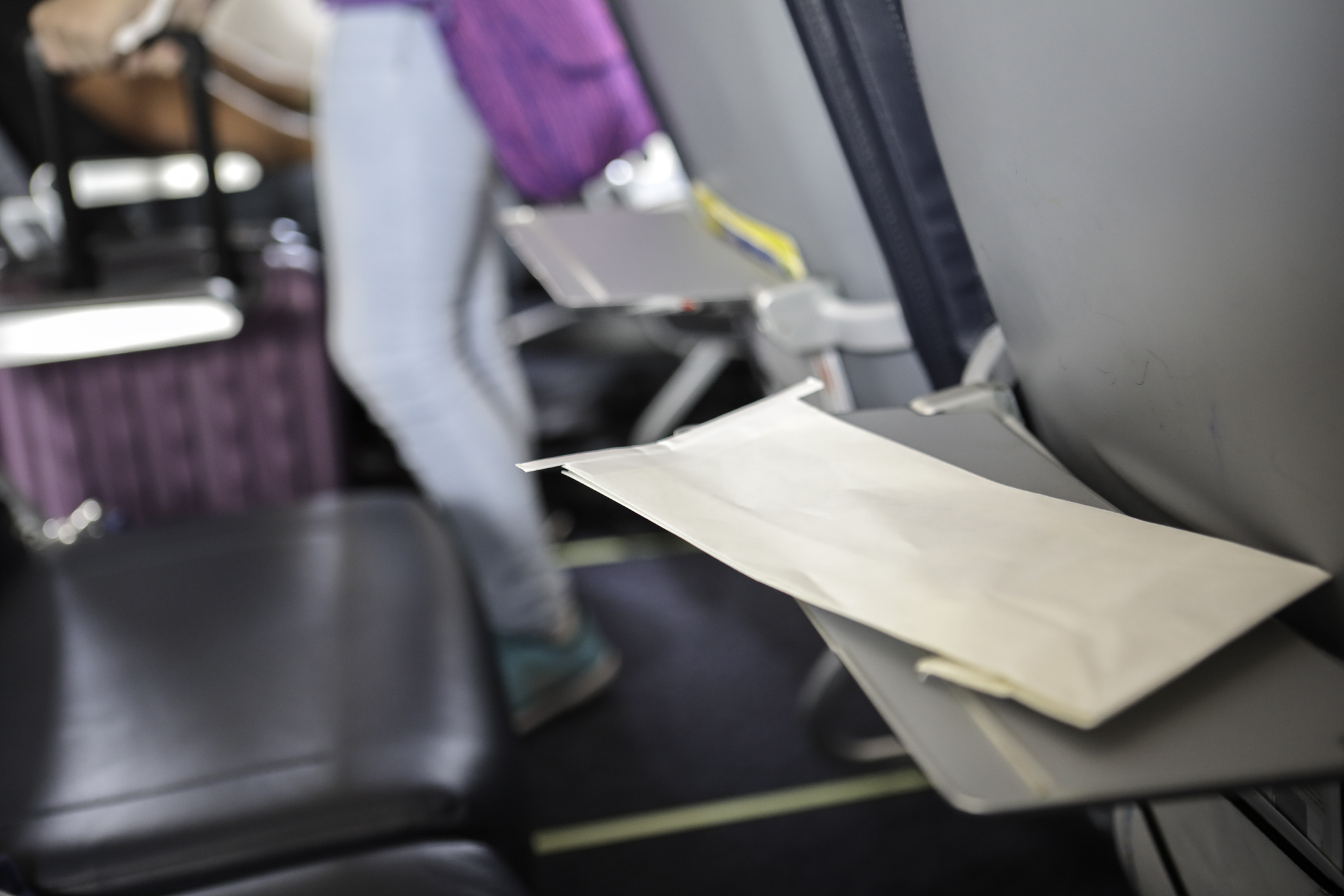 Ein Umschlag liegt auf einem heruntergeklappten Tisch an einem Flugzeugsitz, im Hintergrund sind ein weiterer heruntergeklappter Tisch und eine Person auf dem Gang mit Koffer zu sehen.