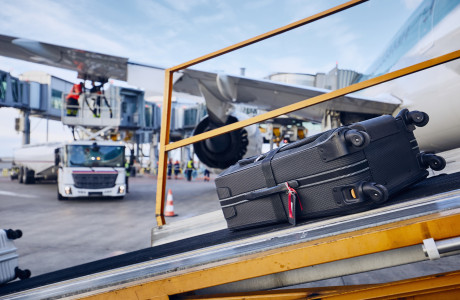 A suitcase is loaded onto an airplane with a luggage belt, in the background the plane is being refueled.