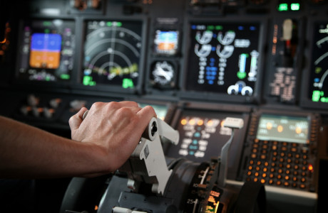 Eine Hand schiebt einen Regler nach vorn, im Hintergrund sind zahlreiche Anzeigen und Messgeräte eines Cockpits zu sehen.