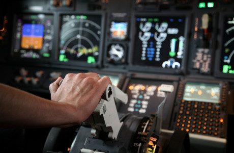 Eine Hand schiebt einen Regler nach vorn, im Hintergrund sind zahlreiche Anzeigen und Messgeräte eines Cockpits zu sehen.