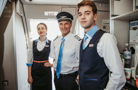 Der Pilot und zwei Flugbegleiter/-innen begrüßen die Passagiere lächelnd.