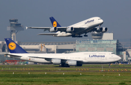 Im Vordergrund steht eine Lufthansa-Maschine auf dem Runway eines Flughafens, im Hintergrund hebt eine zweite gerade ab.