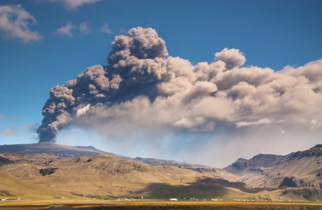 Op de voorgrond zijn enkele huizen in IJsland te zien, met Eyjafjallajökull en zijn aswolk op de achtergrond.